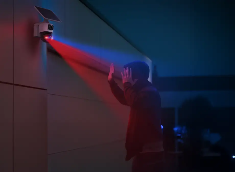 Cámara de seguridad eufyCam S4 activando luces de advertencia rojas y azules por la noche ante una persona con capucha junto a la pared exterior