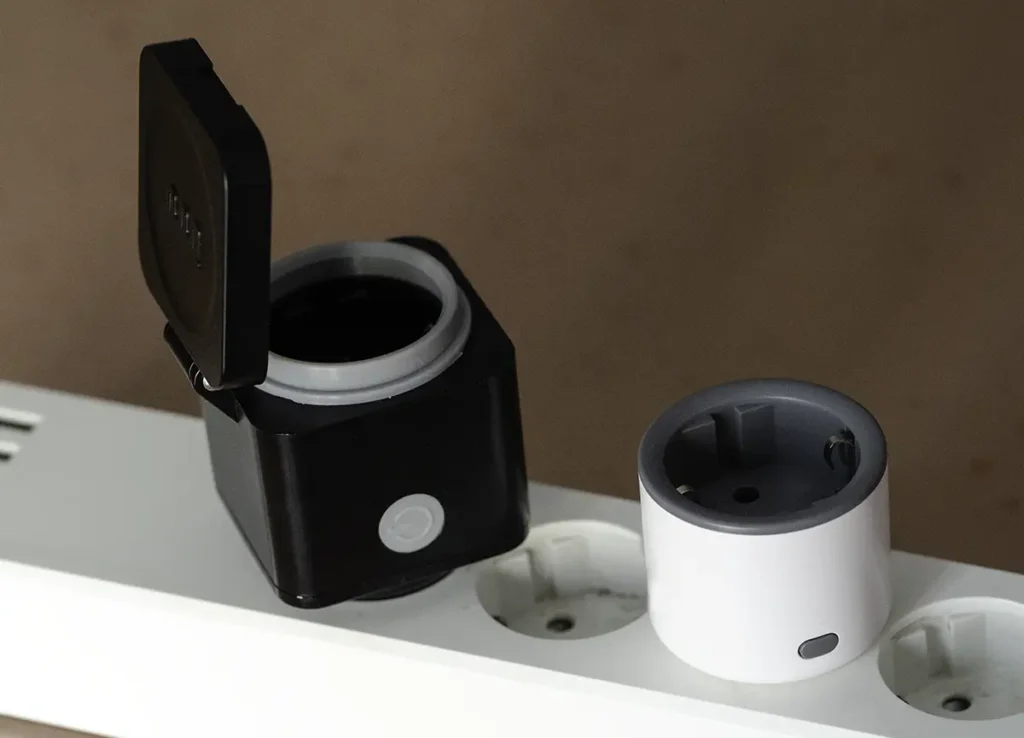 Enchufe exterior Innr OSP 240 con tapa abierta junto a un enchufe Sonoff S60ZBTPF, ambos colocados en una regleta para comparar tamaño