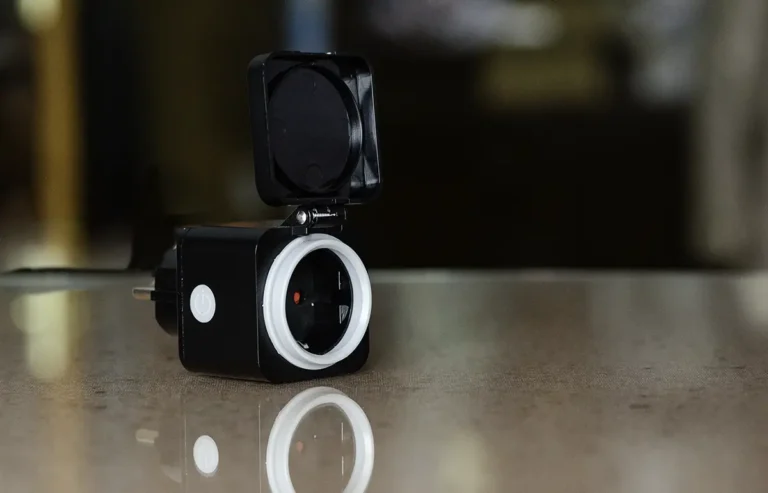 Enchufe inteligente exterior Innr OSP 240 con tapa abatible y botón lateral, colocado sobre una mesa y fotografiado en primer plano con fondo desenfocado.