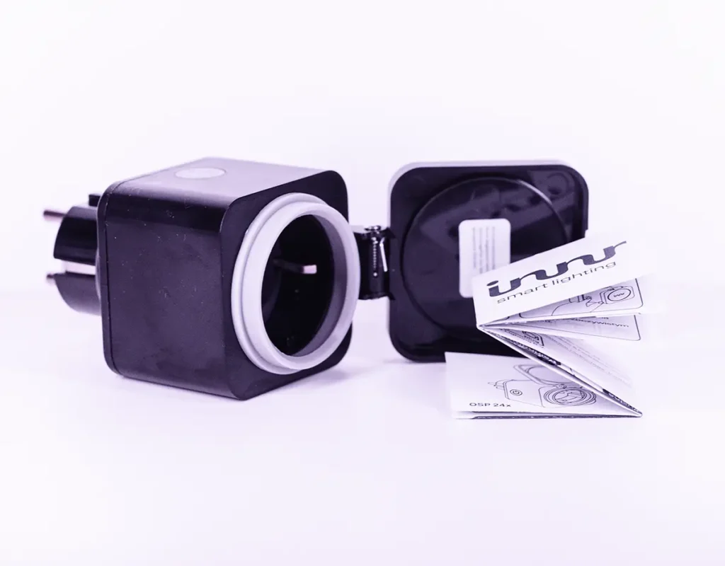 Enchufe inteligente exterior Innr OSP 240 con tapa abatible abierta junto al manual de usuario, fotografiado sobre fondo blanco.