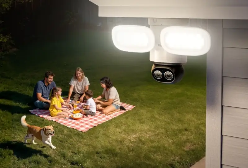 Reolink TrackFlex Floodlight WiFi iluminando un jardín por la noche mientras una familia está sentada sobre una manta y un perro corre cerca