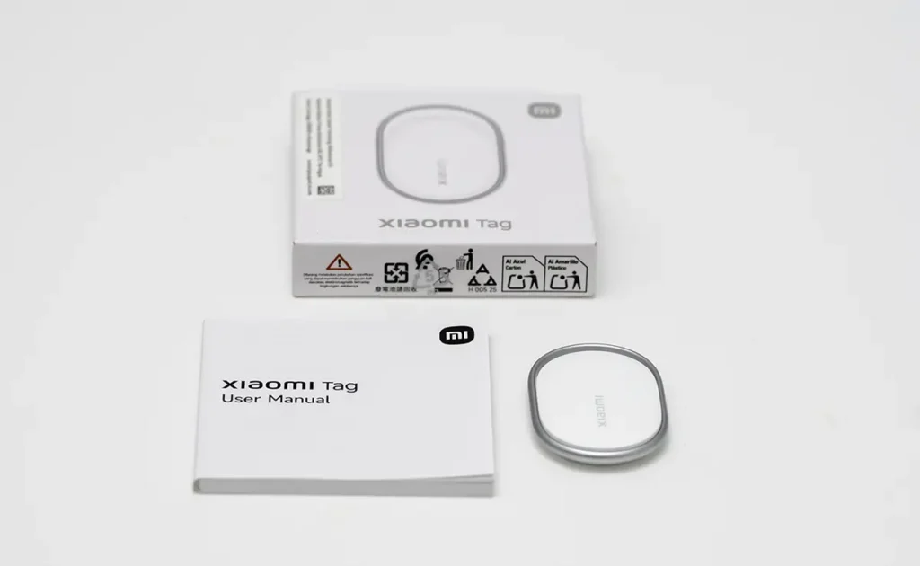 Unboxing del Xiaomi Tag con la caja, el manual de usuario y el rastreador sobre fondo blanco.