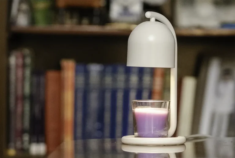 SwitchBot Candle Warmer Lamp blanco calentando una vela aromática morada sobre una mesa, con una estantería desenfocada al fondo