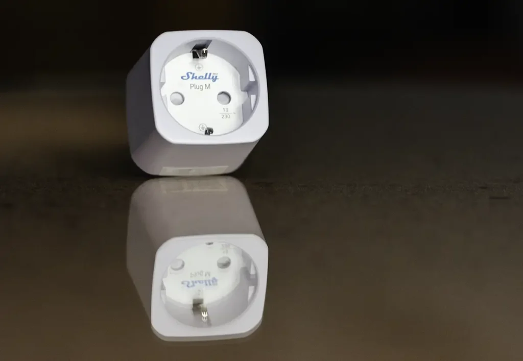 Shelly Plug M Gen3 sobre una superficie reflectante, mostrando el diseño cuadrado del enchufe inteligente con Matter y medición de consumo.
