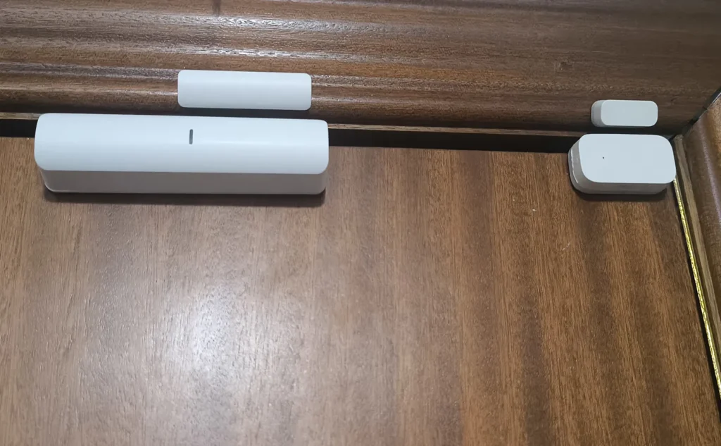 Sensor de puerta y ventana ANNKE instalado en una puerta de madera junto a un sensor Aqara para comparar tamaño y colocación.